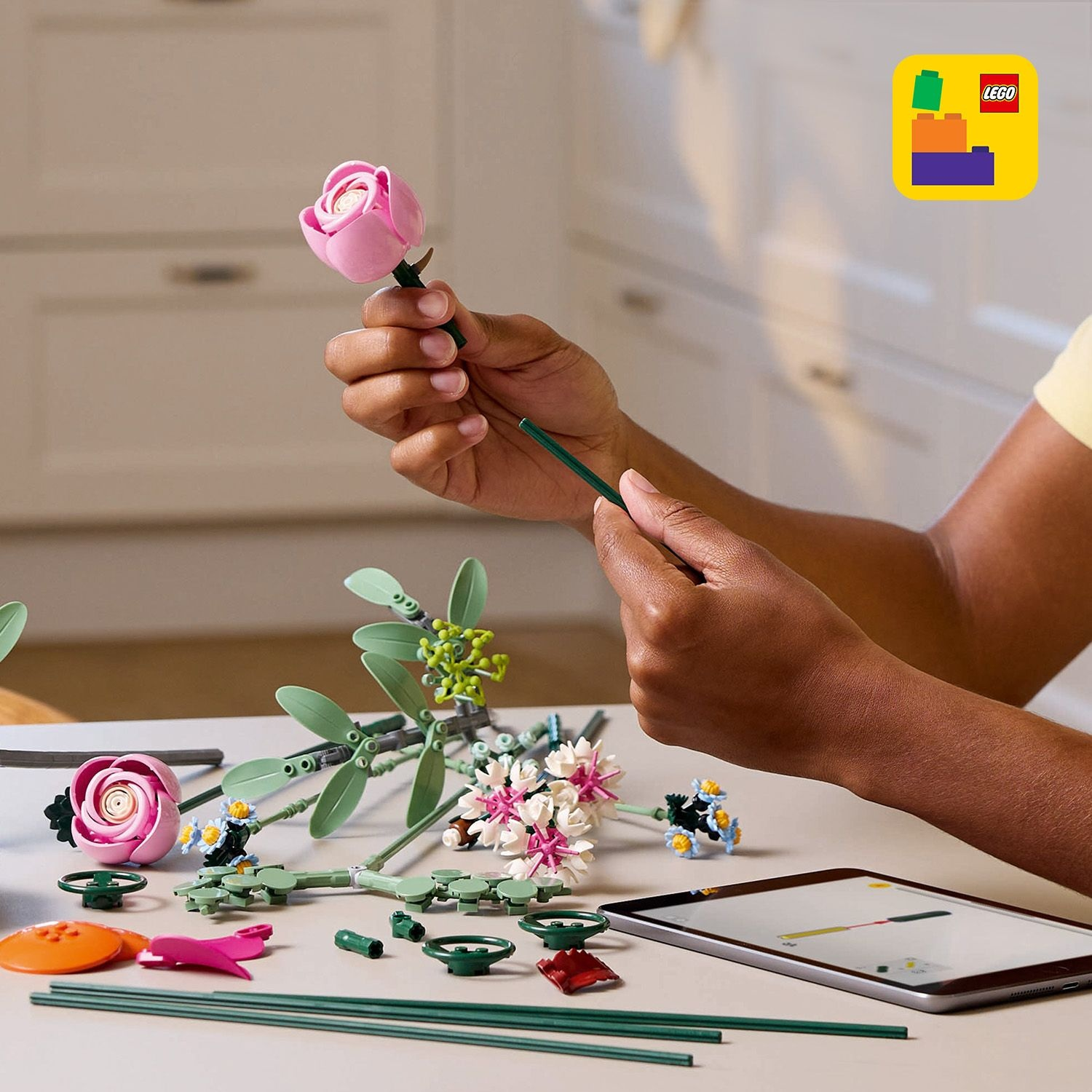 Mani che assemblano fiori Lego. Rosa rosa tenuta, steli e foglie verdi, altri fiori sul tavolo, istruzioni su un tablet e logo Lego.