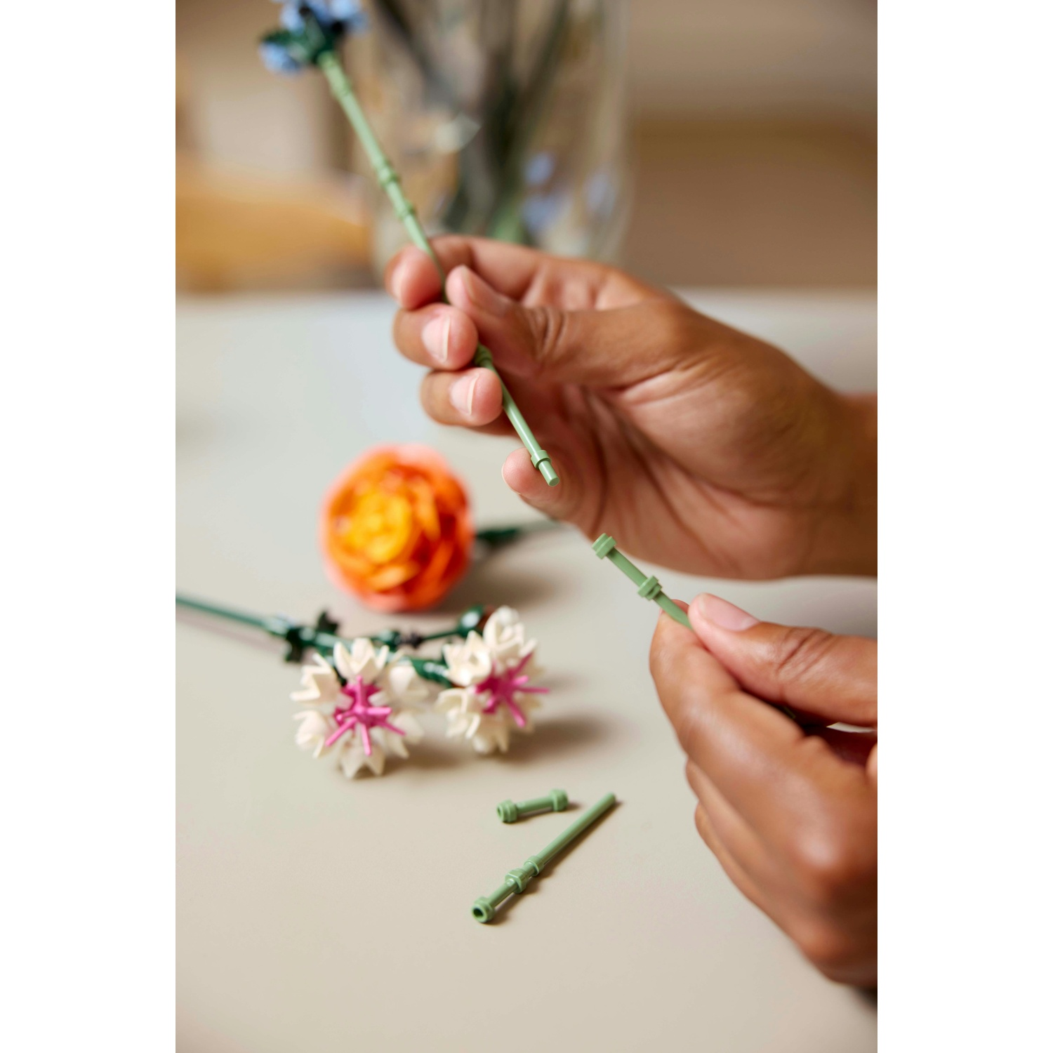 Mani che assemblano fiori LEGO. Una mano tiene uno stelo, l'altra collega un pezzo verde. Altri fiori sono sul tavolo.