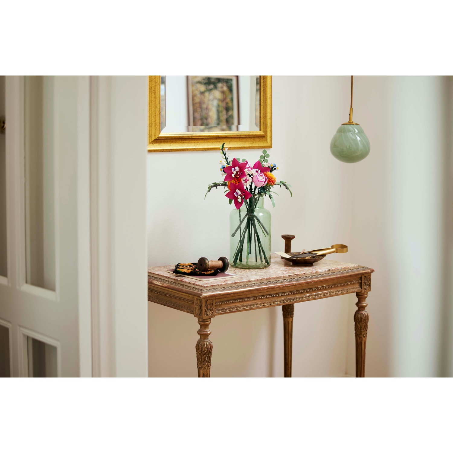 Un tavolo con fiori in un vaso, uno specchio e una lampada a sospensione. Il tavolo ha gambe ornate e un piano in marmo.