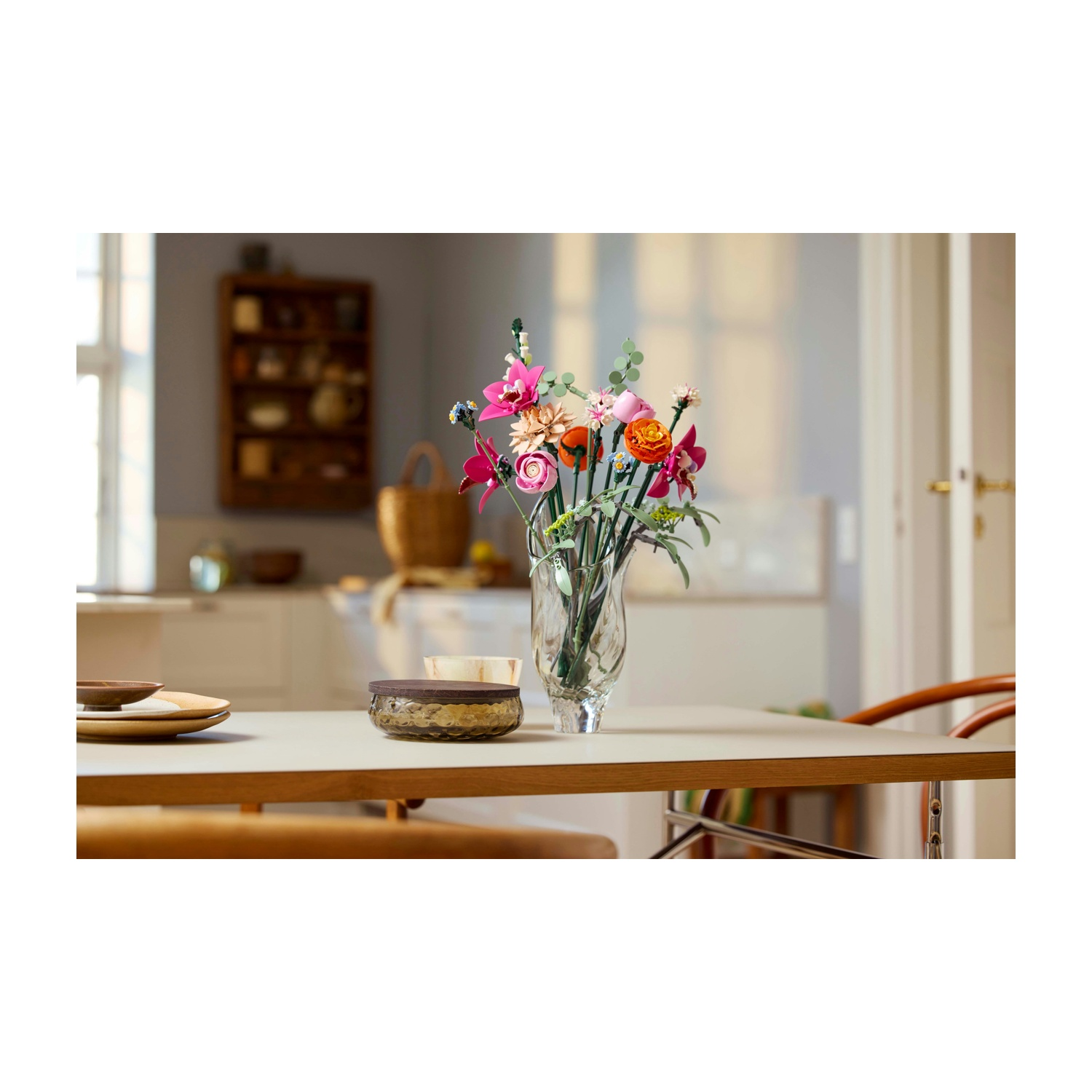 Un tavolo con un vaso di fiori. Sono visibili anche una ciotola, dei piatti e delle sedie. Sullo sfondo una mensola e una porta.