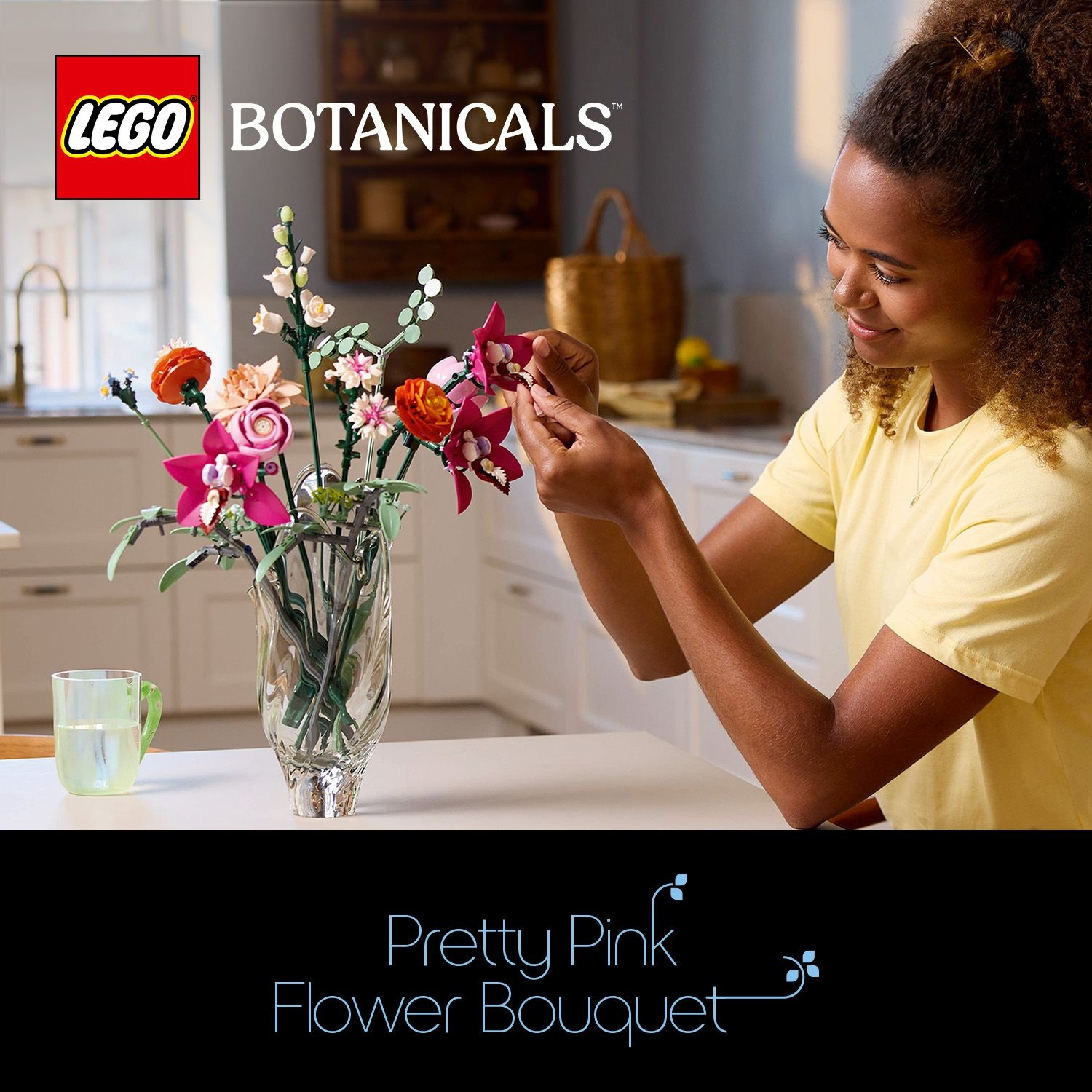 Una donna sistema un mazzo di fiori Lego. Il bouquet contiene fiori rosa, arancioni e bianchi in un vaso. Lo sfondo è una cucina.