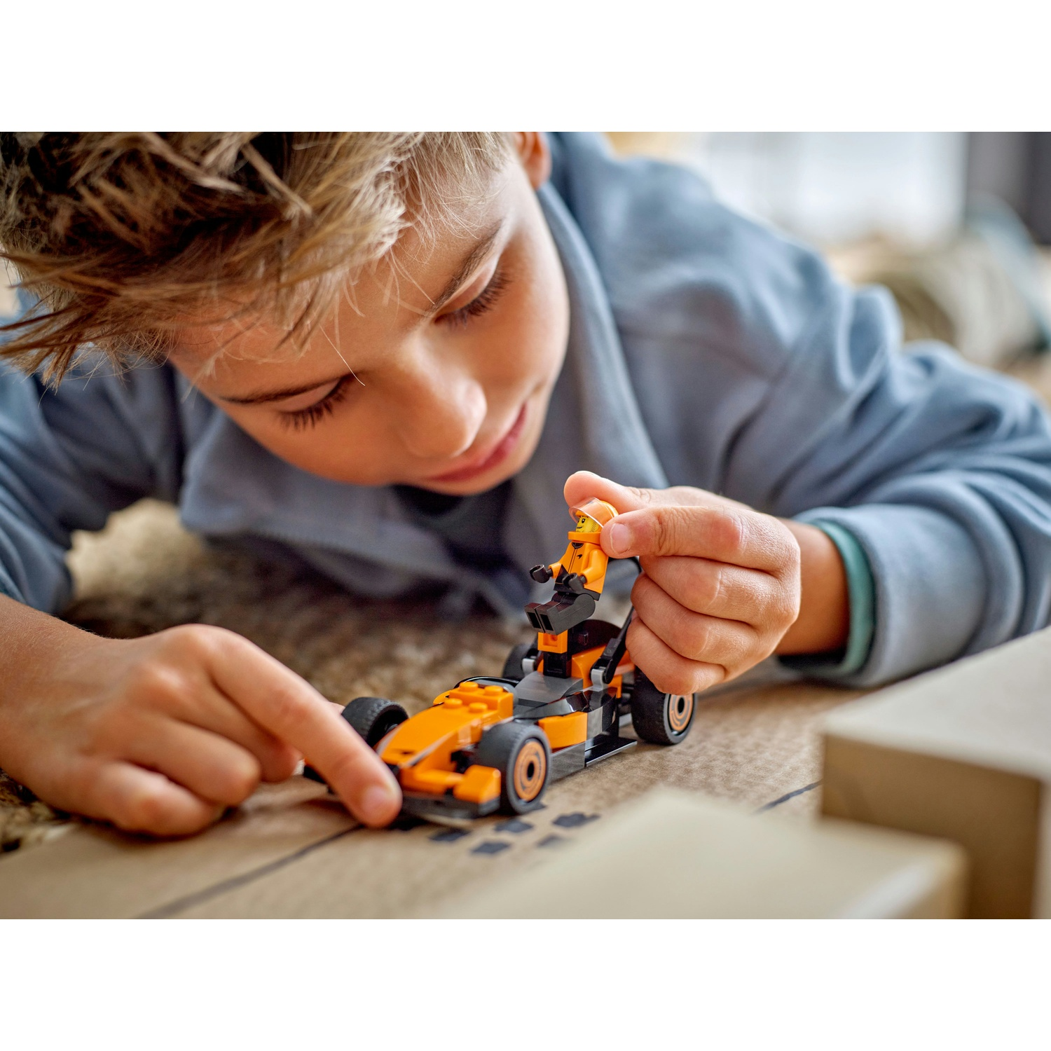 Un bambino esamina un'auto da corsa Lego arancione e nera. L'auto è su una superficie marrone. Il bambino indossa una camicia blu.