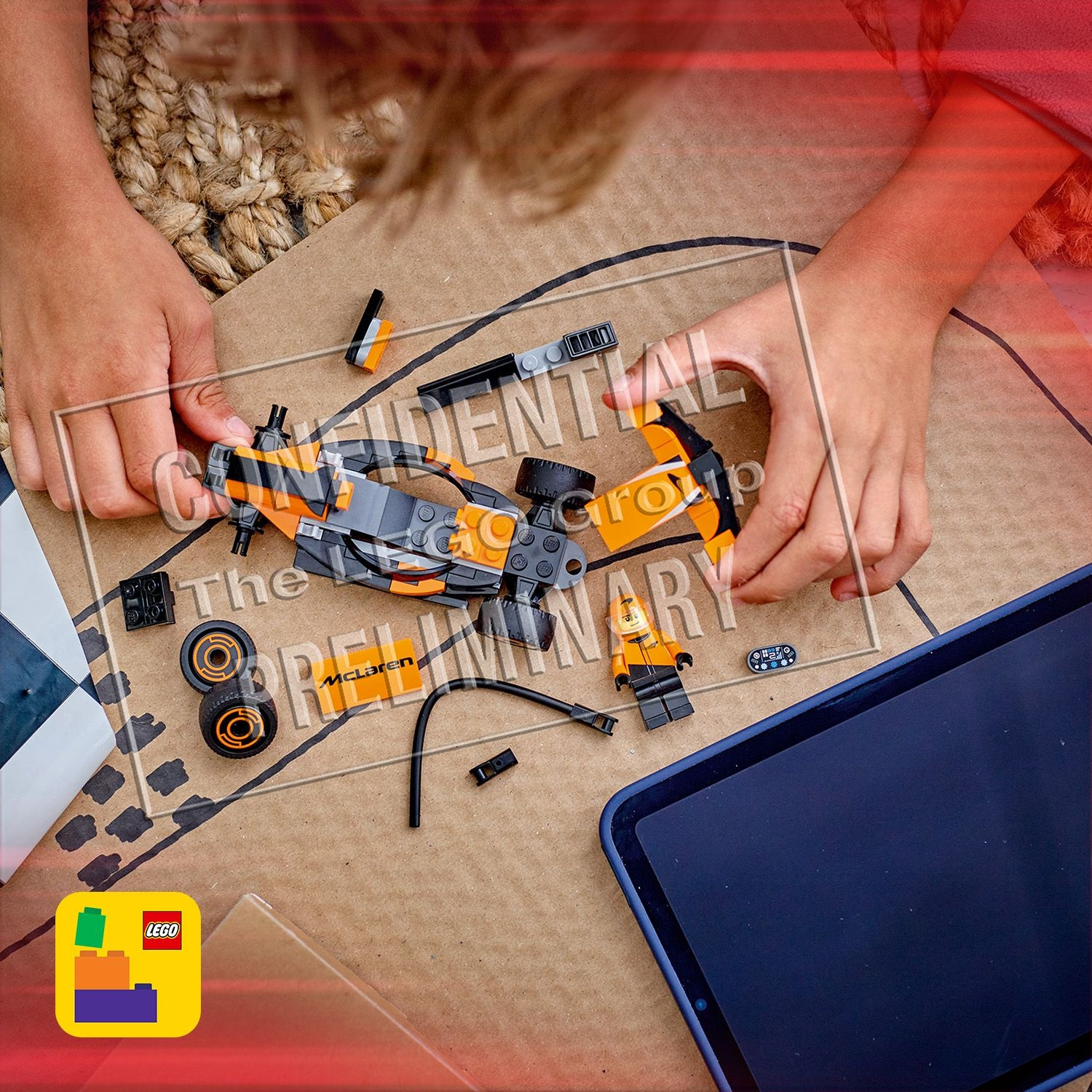Mani che assemblano un'auto da corsa Lego McLaren su una superficie di cartone. Parti e un tablet sono vicini.