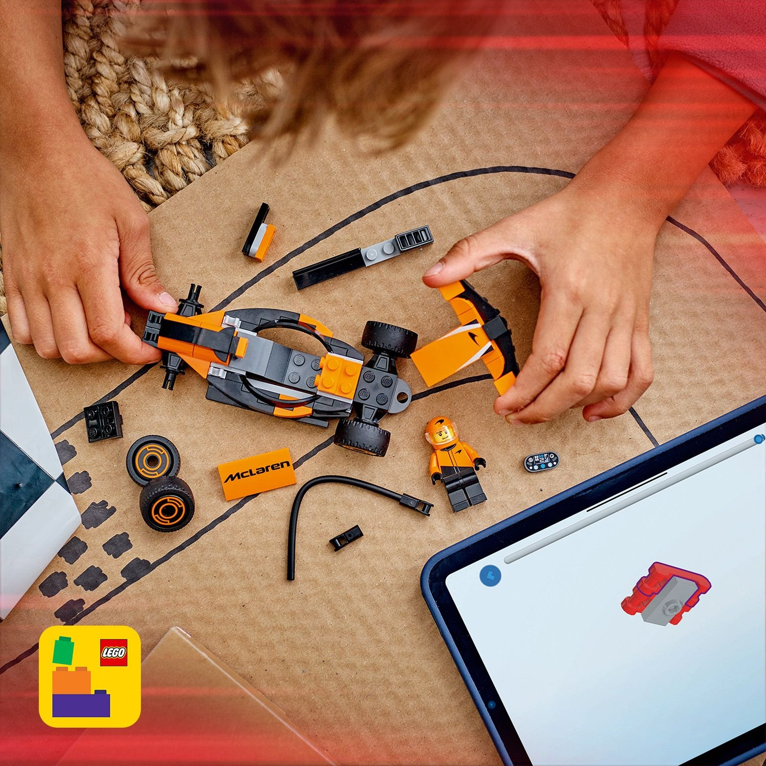 Mani che assemblano un'auto da corsa McLaren Lego. Parti e istruzioni visibili. Un tablet mostra i passaggi di costruzione. Lo sfondo è una superficie marrone.