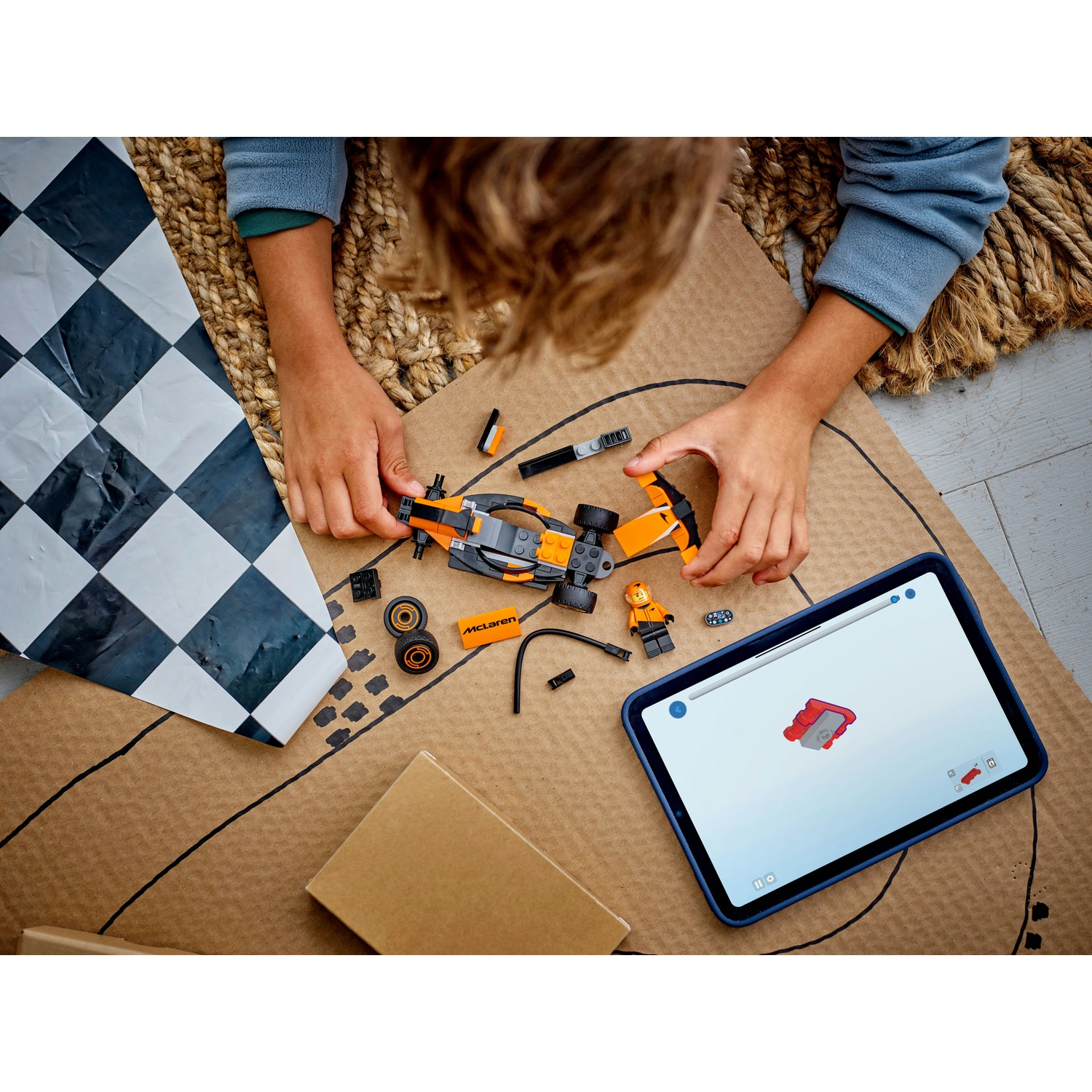 Un bambino assembla un'auto da corsa Lego, usando un tablet per le istruzioni. I pezzi sono sparsi su cartone, con una bandiera a scacchi visibile.