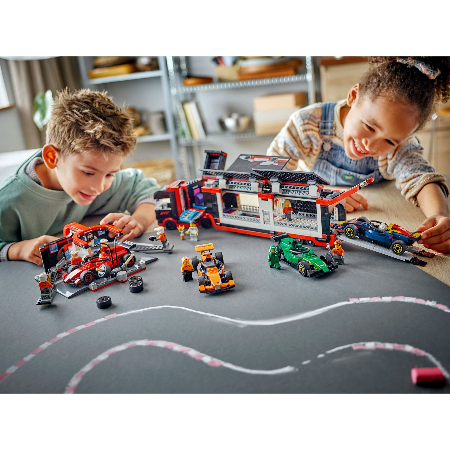 Due bambini giocano con auto da corsa LEGO e un garage su una superficie grigia con una pista disegnata.