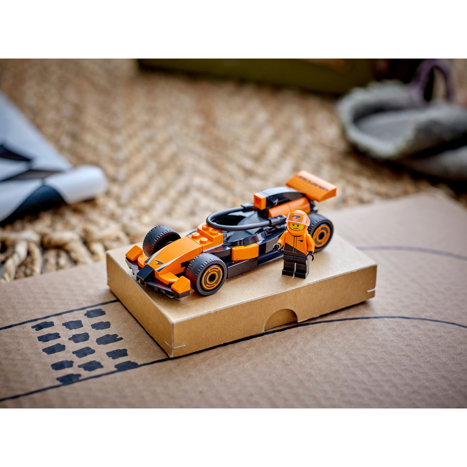 Un'auto da corsa Lego e una minifigure sono su una scatola di cartone. L'auto è arancione e nera. Lo sfondo ha un tappeto e altri oggetti.
