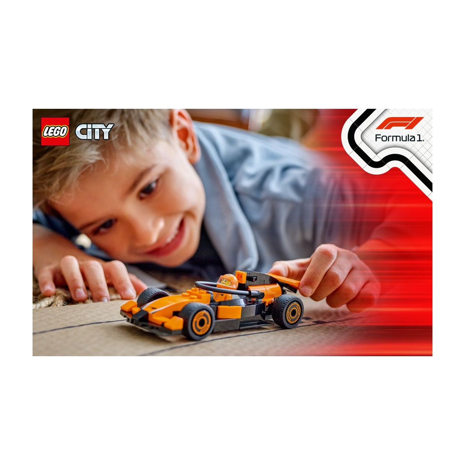 Un ragazzo sorride, guardando una macchina da corsa Lego arancione e nera. L'auto è su una superficie marrone. Logo Formula 1 a destra.