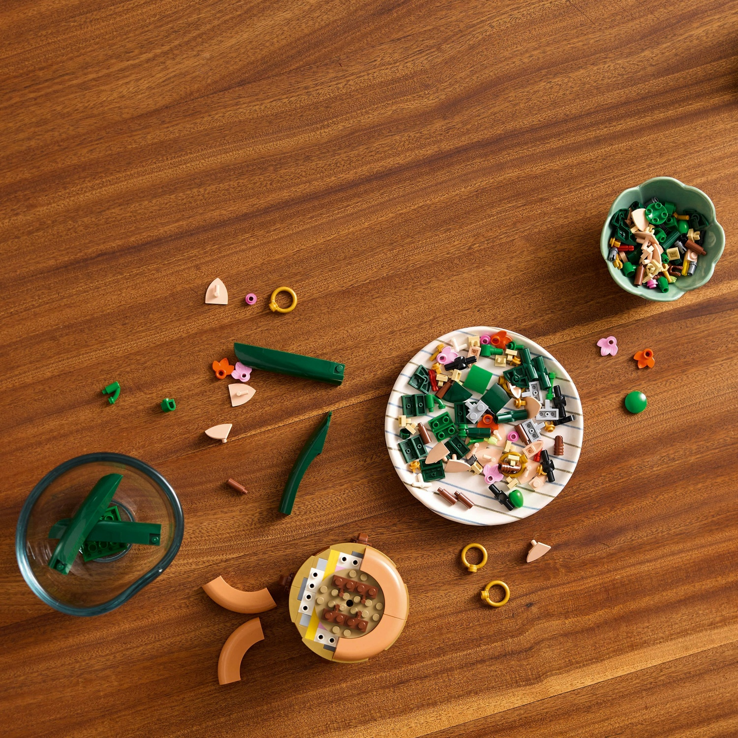 Tavolo di legno con pezzi Lego sparsi: verde, beige, rosa e oro. Ciotole con Lego e una struttura circolare parzialmente assemblata.
