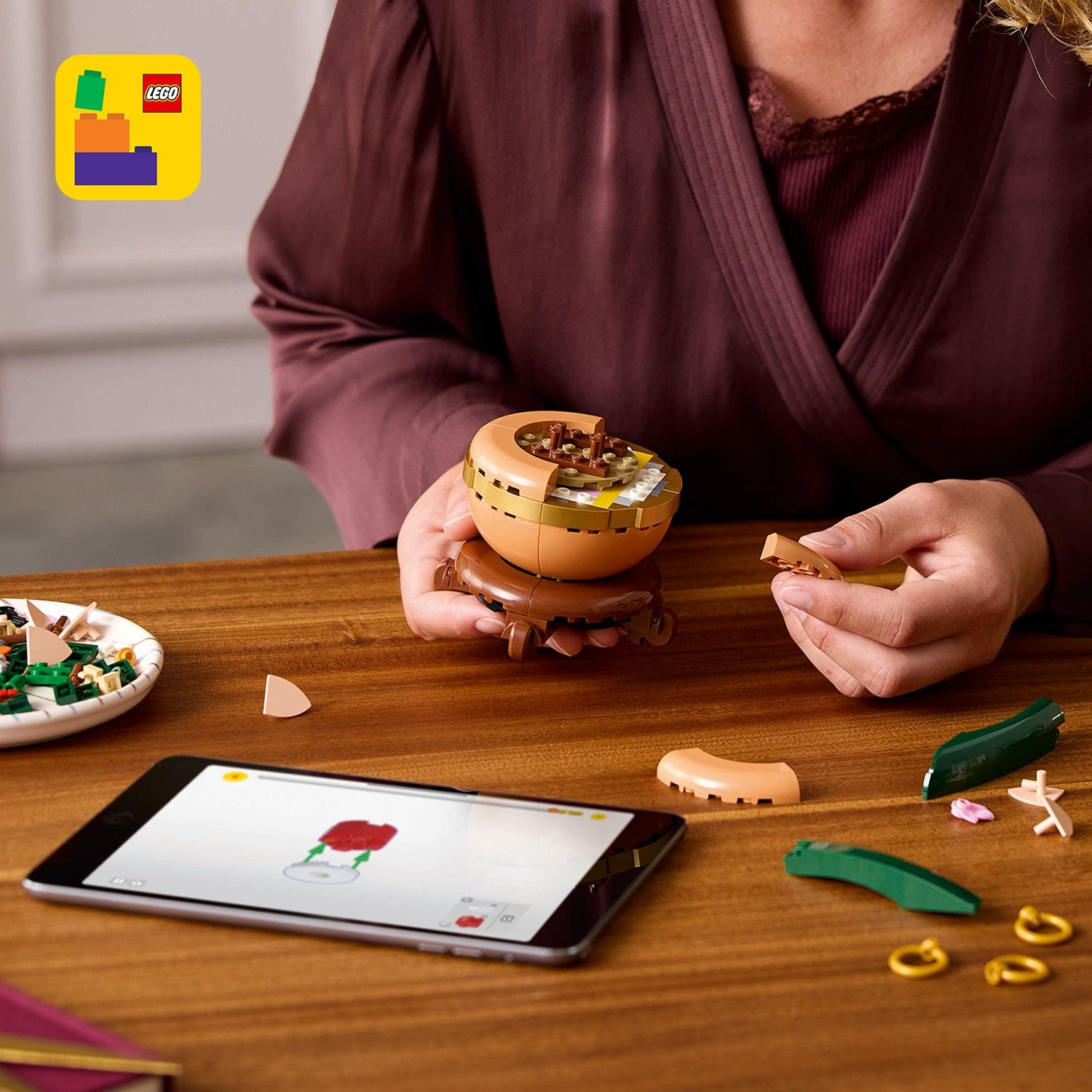 Una persona assembla un set Lego. Il set è su un tavolo di legno. Un tablet mostra le istruzioni. Vari pezzi Lego sono sparsi.