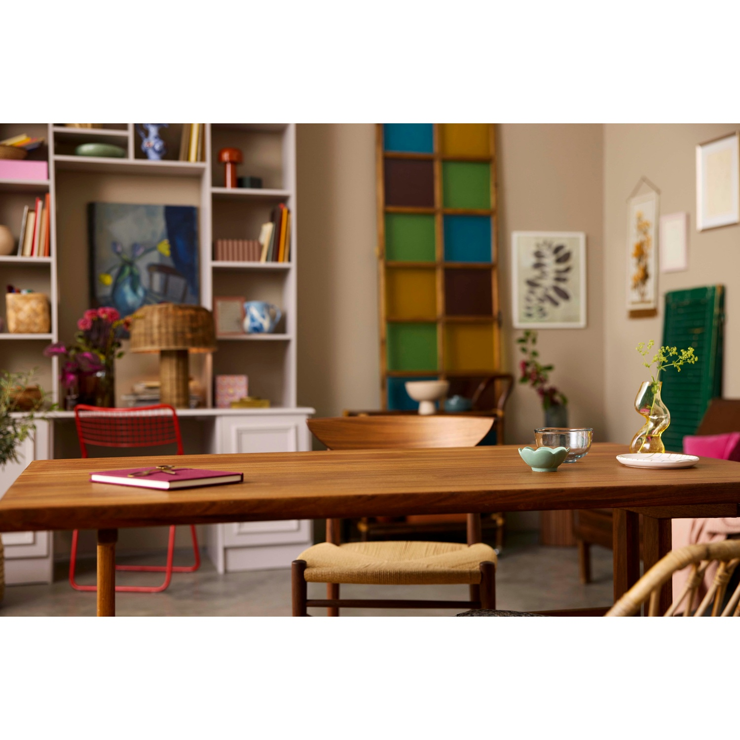 Tavolo in legno con libro, ciotola e bicchiere. Sullo sfondo, una stanza con scaffali, opere d'arte e una finestra colorata.