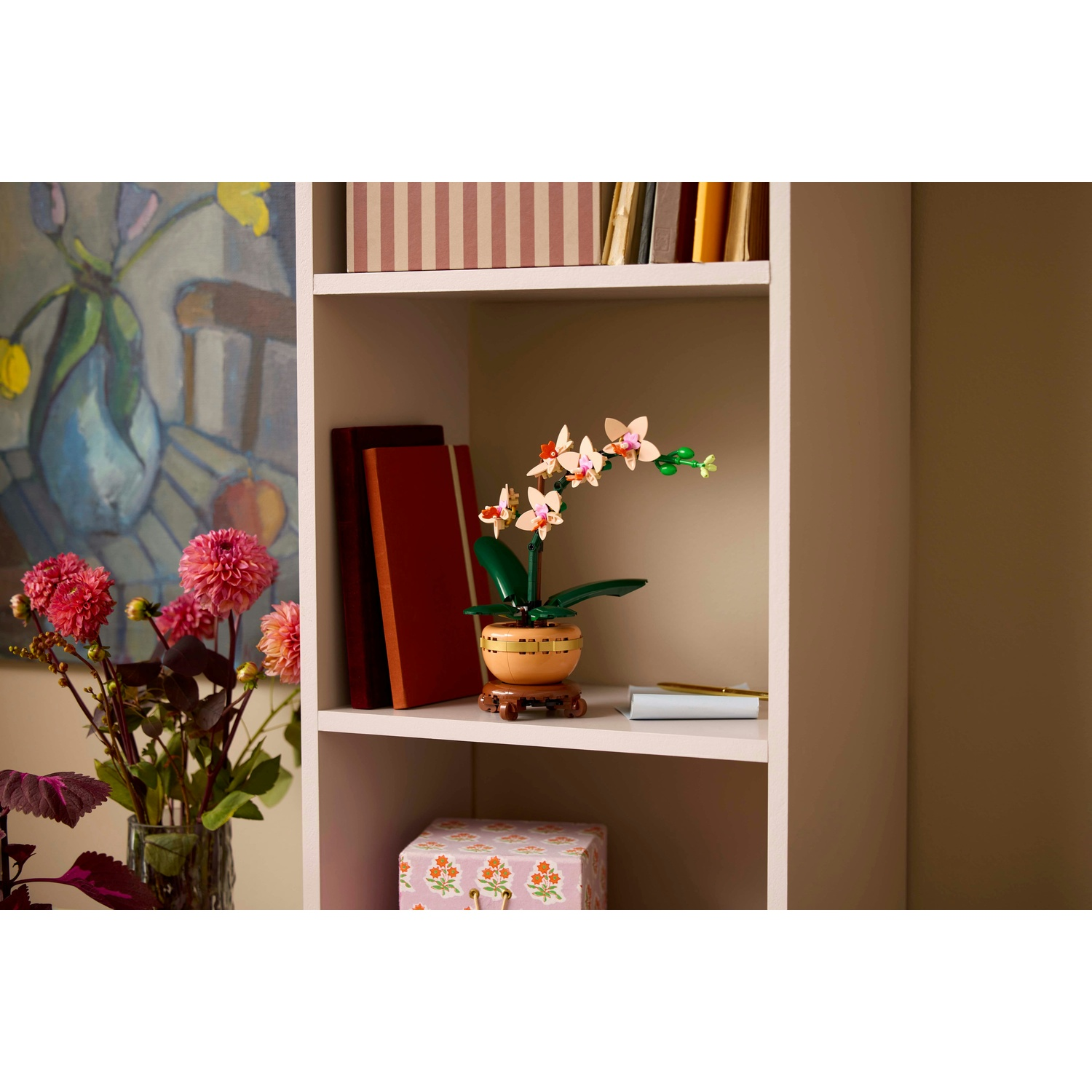 Una mensola bianca con un'orchidea Lego, libri e una scatola floreale. Sono presenti anche un vaso di fiori rosa e un dipinto.