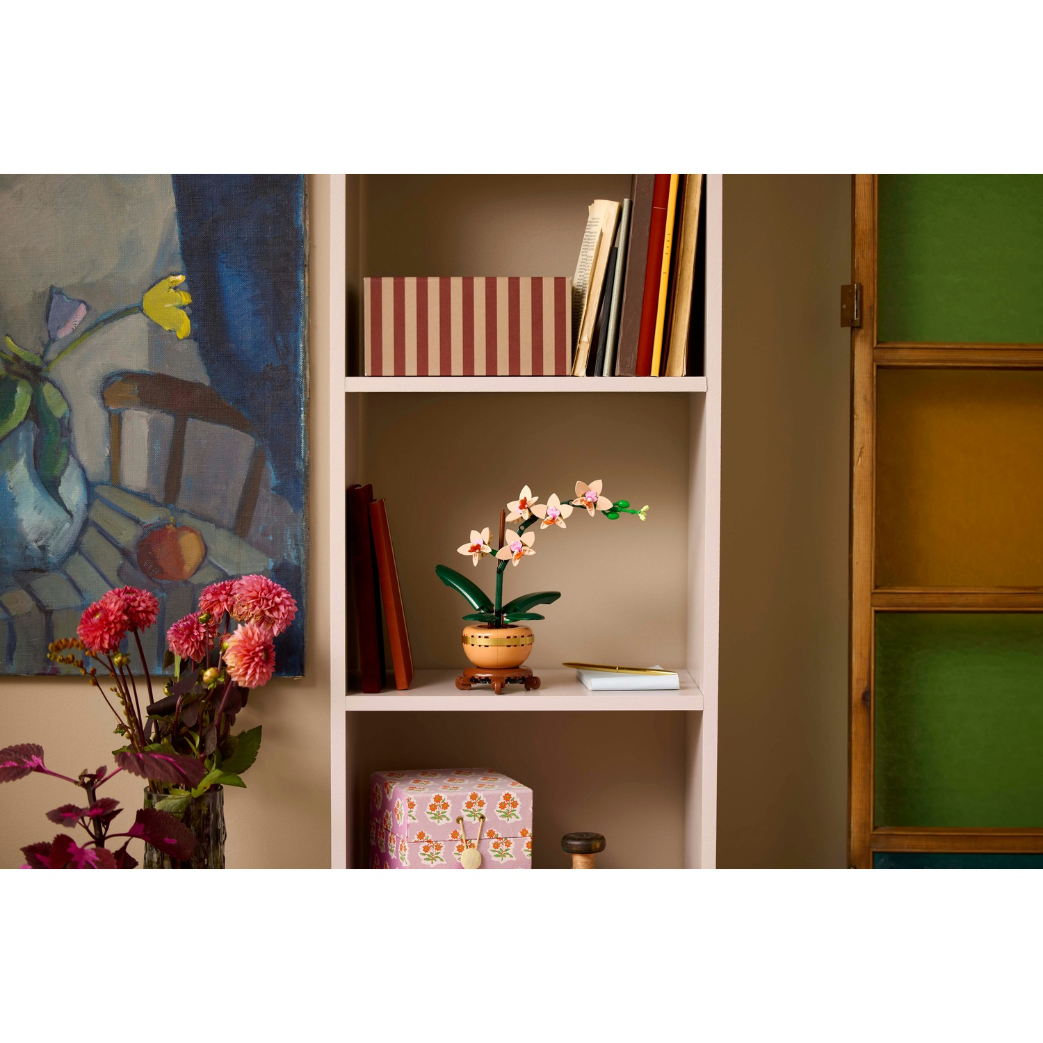 Una mensola mostra un'orchidea, libri e una scatola. Un vaso con fiori rosa è a sinistra. Sono visibili un dipinto e una porta a vetri colorati.