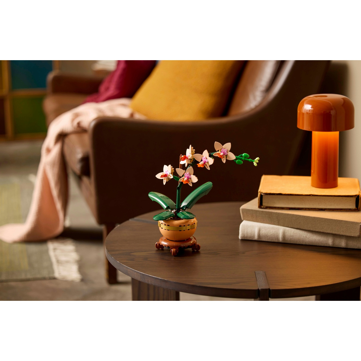 Un'orchidea Lego in vaso su un tavolo. Accanto una lampada e libri. Sullo sfondo un divano con cuscini.