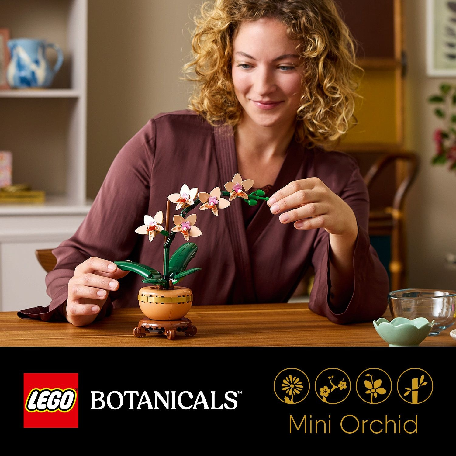 Donna con i capelli ricci esamina il set di orchidee LEGO. L'orchidea ha fiori rosa e bianchi, in un piccolo vaso su un supporto di legno.