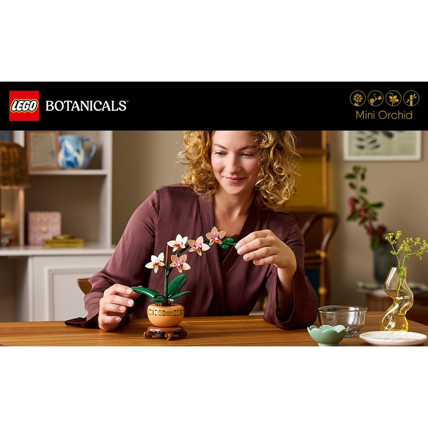 Donna con orchidea LEGO sul tavolo. Sorride. Altri oggetti: ciotola, piatto e vaso con fiori.