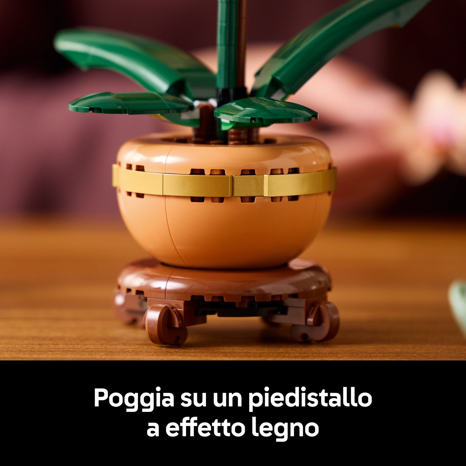Una pianta Lego in un vaso su un supporto effetto legno. Foglie verdi spuntano dal vaso. Il vaso è marrone, con una fascia dorata.