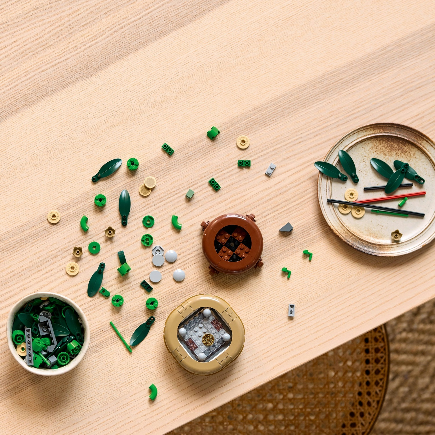 Tavolo di legno con pezzi Lego sparsi. Include foglie verdi, elementi circolari marroni e beige e due strutture Lego. È presente un piatto.