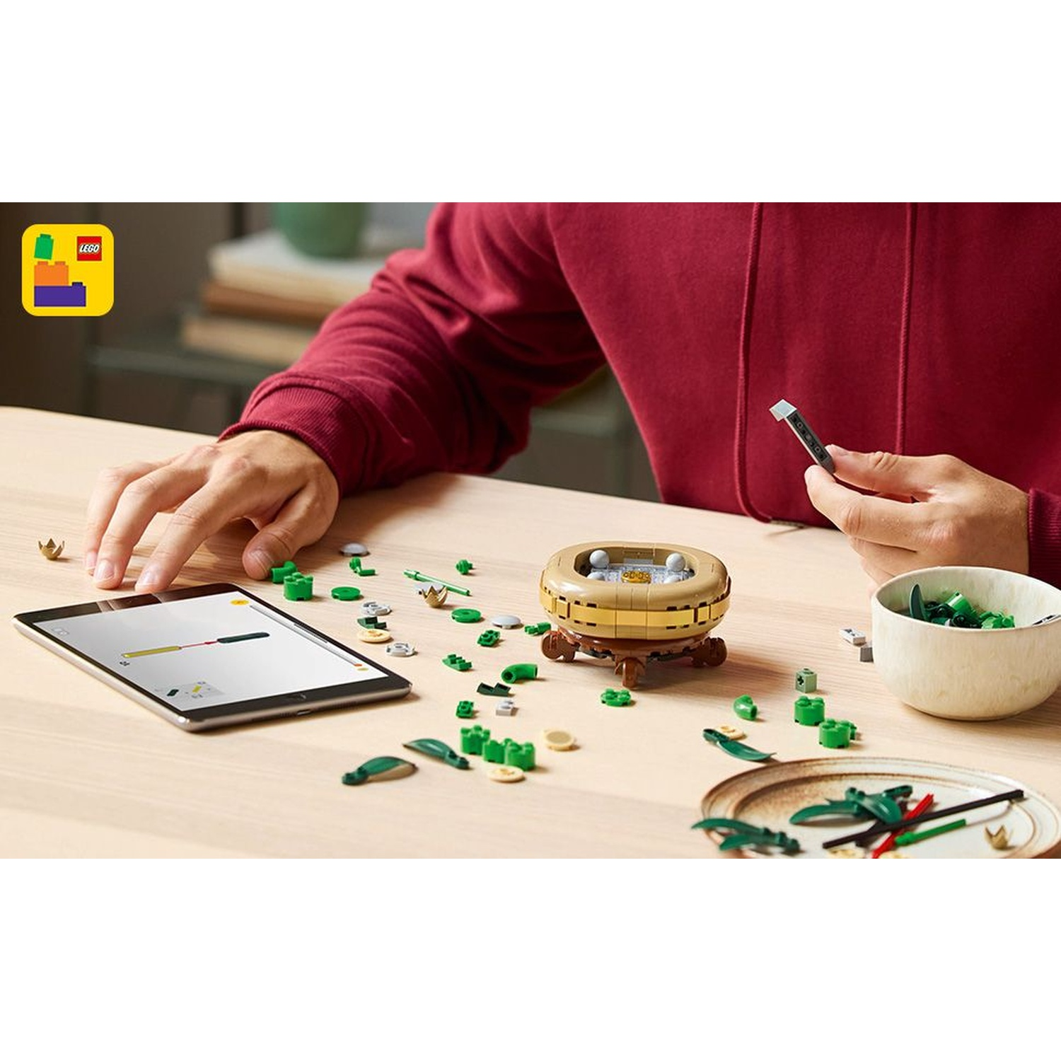 Una persona assembla un set Lego. La scena include un tablet, vari pezzi Lego, una ciotola e il modello parzialmente costruito.