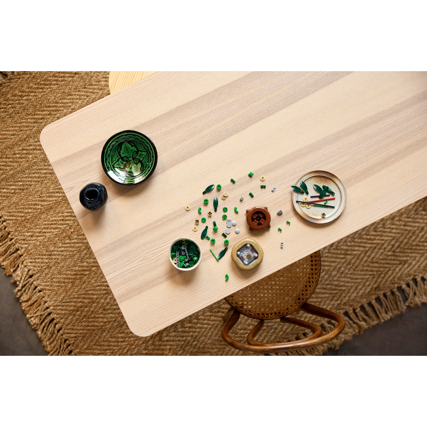 Vista dall'alto di un tavolo di legno con oggetti: ciotola verde, vaso nero, piatti con foglie e una sedia in vimini.