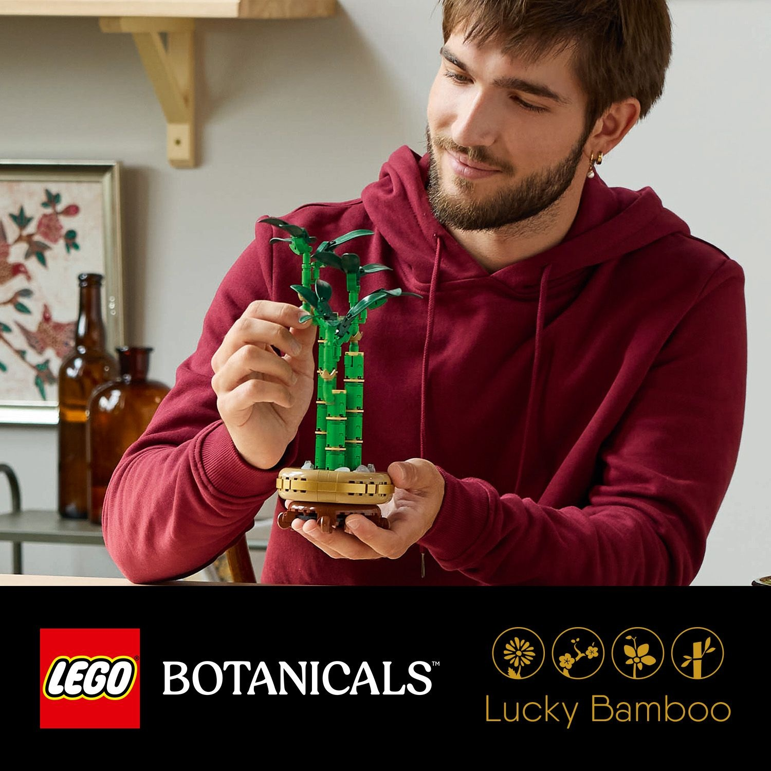Un uomo con una felpa rossa tiene un Lucky Bamboo LEGO. Il set è verde, marrone e oro. Sfondo: mensola, bottiglie e dipinto.