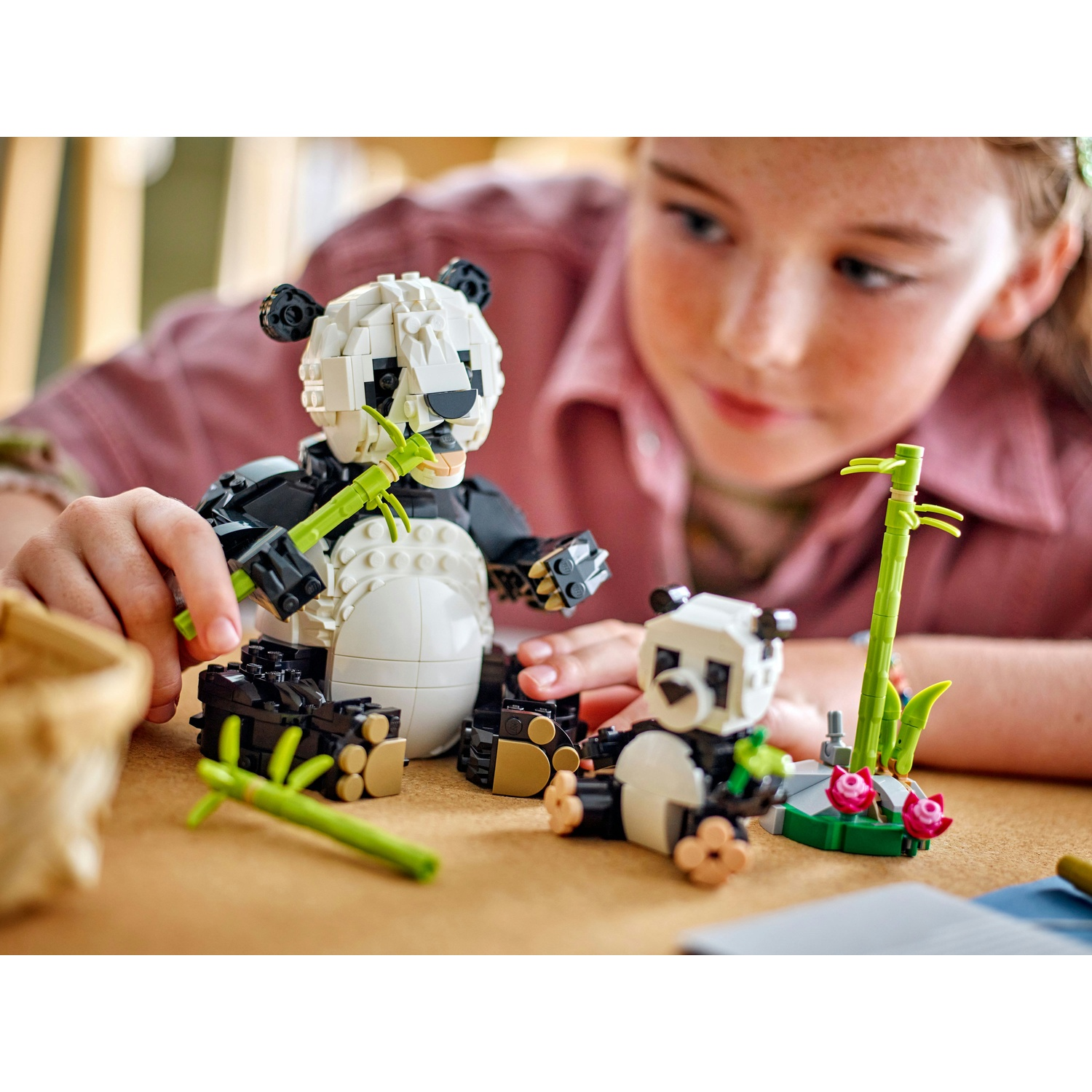 Una ragazza gioca con set Lego di panda. Un grande panda mangia bambù, un altro piccolo è lì vicino, e ci sono bambù e fiori.