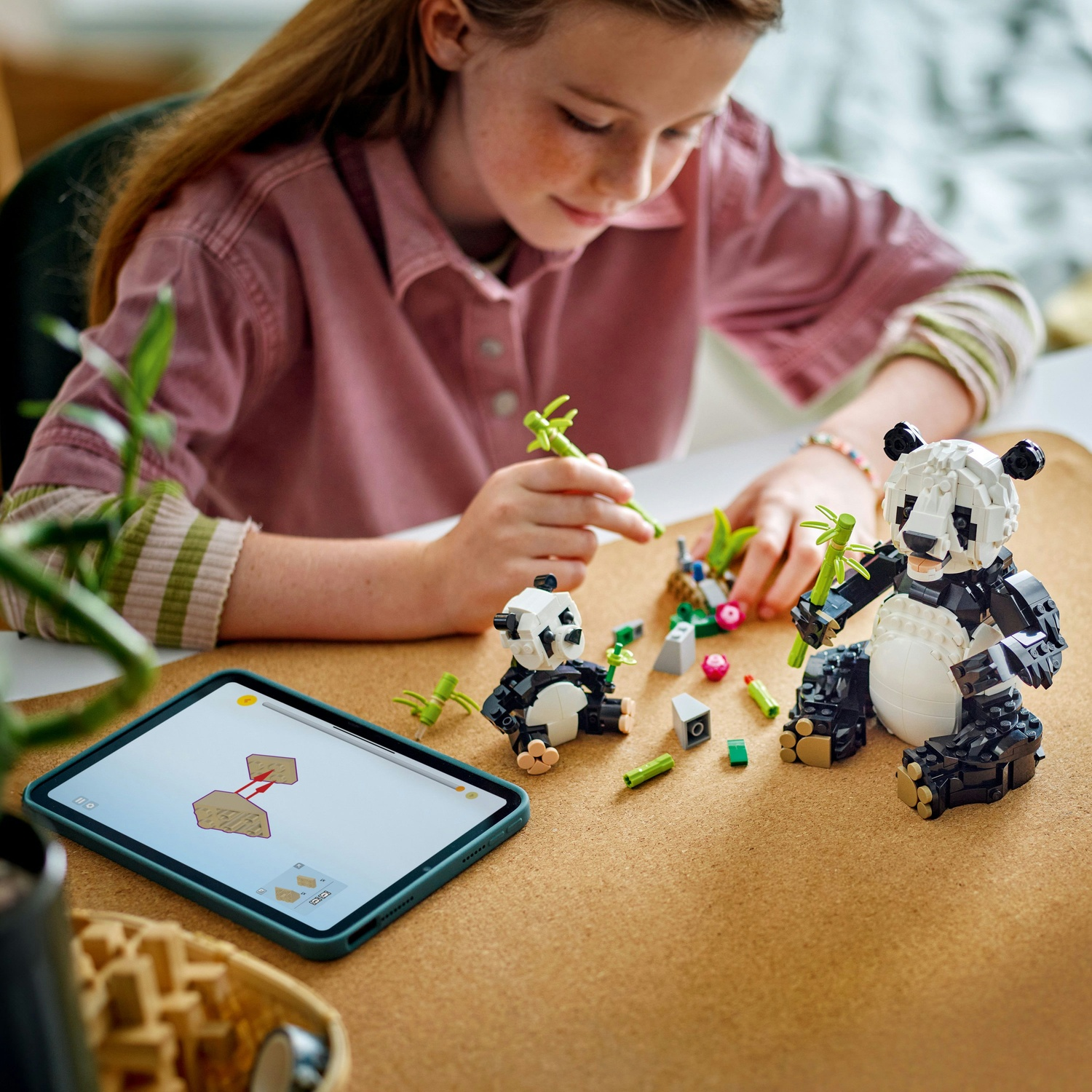 Una ragazza costruisce set Lego di panda su una superficie marrone. Un tablet mostra le istruzioni. Bambù e altri pezzi Lego sono sparsi.