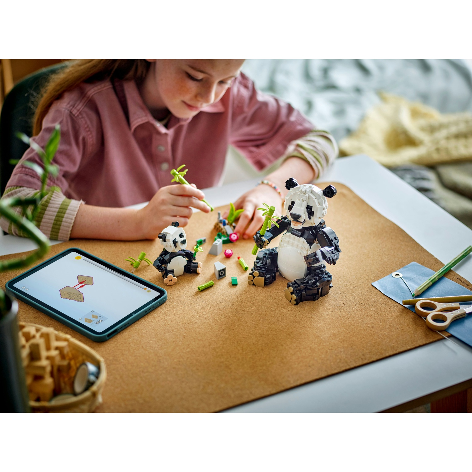 Una ragazza costruisce un panda Lego. Ci sono due panda, bambù e un tablet con le istruzioni su un tavolo di legno.