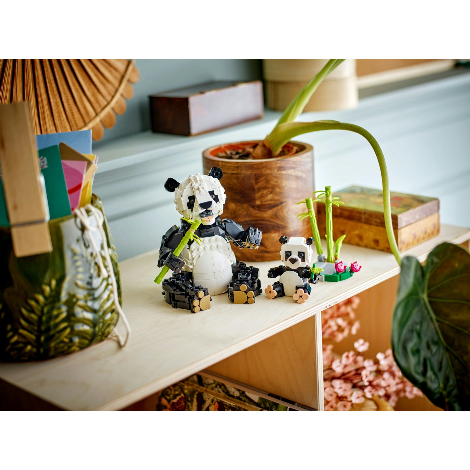 Due panda Lego seduti su una mensola. Uno grande, con bambù, l'altro piccolo. Dietro, una pianta in vaso di legno e altri oggetti decorativi.