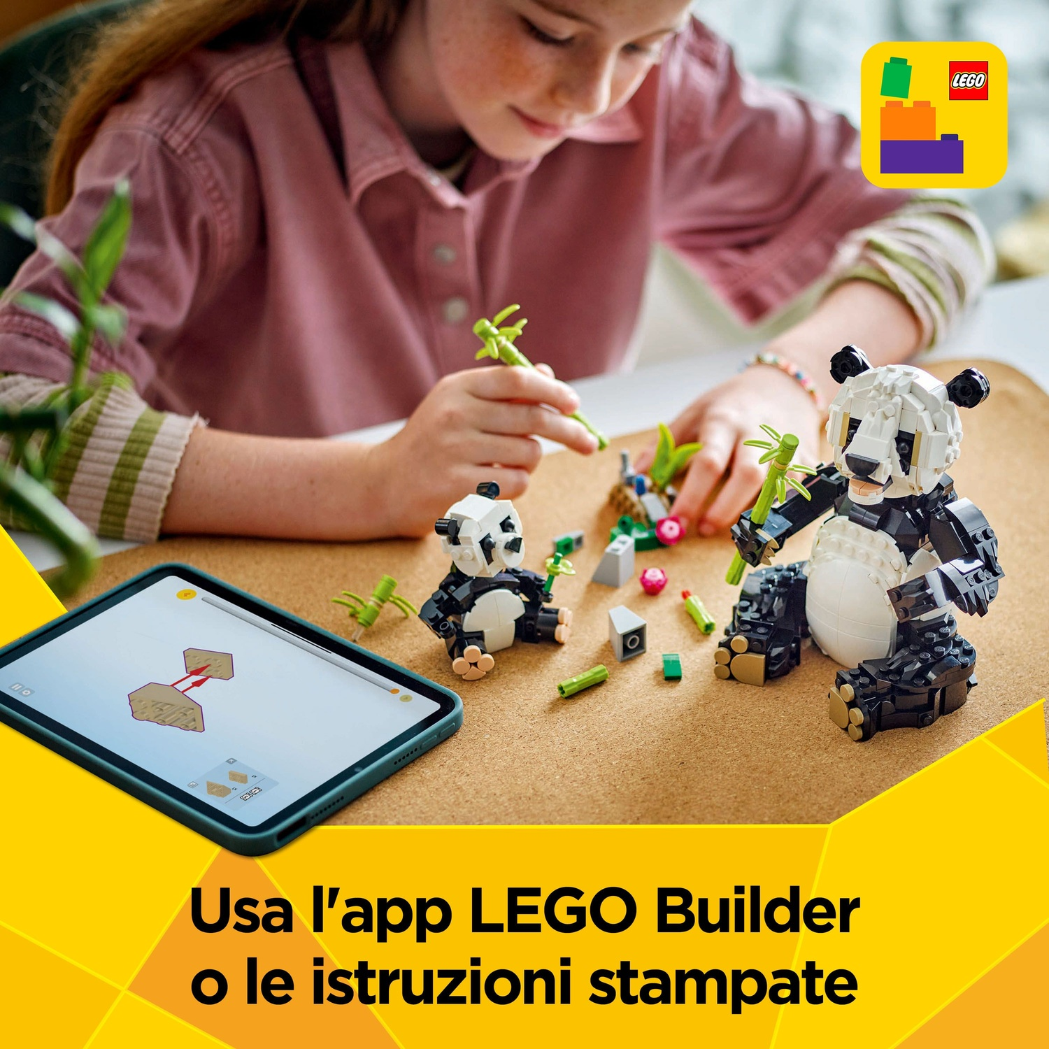 Una ragazza costruisce un panda Lego. Un tablet mostra le istruzioni. Due panda Lego su una tavola di sughero. Pezzi Lego verdi sparsi.
