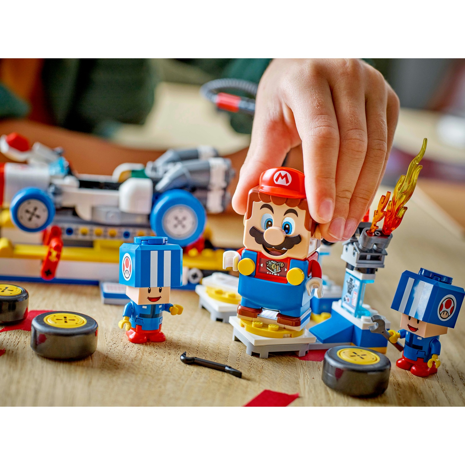 Una mano posiziona un personaggio Lego di Mario. Il set Lego ha Mario, due personaggi blu, un'auto, pneumatici e una struttura a fiamma, il tutto su una superficie di legno.