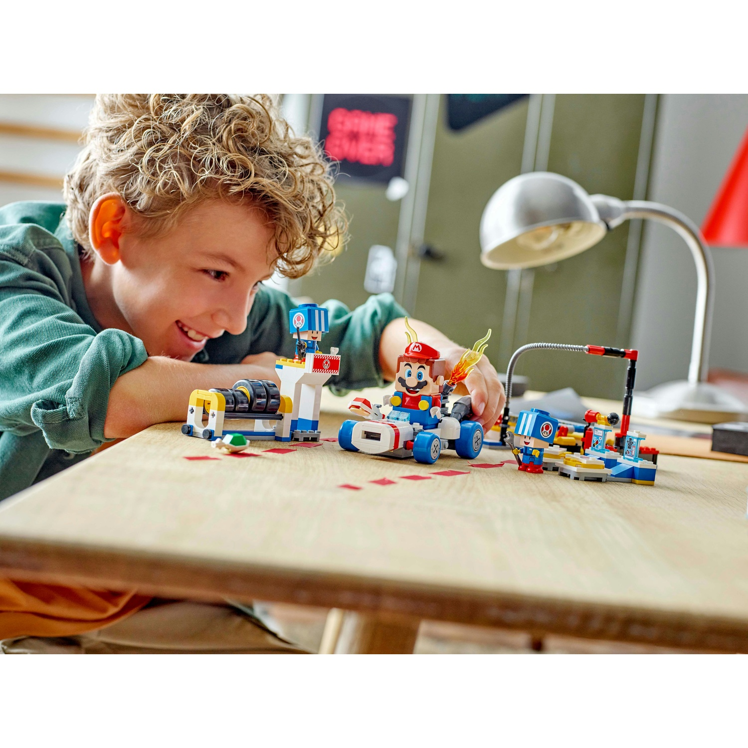 Un ragazzo sorride mentre gioca con un set Lego di Mario. Il set include personaggi e veicoli su un tavolo di legno.