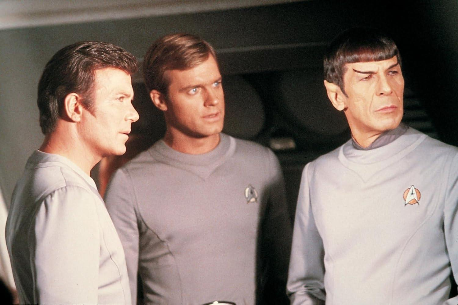 Drei Männer in grauen Star-Trek-Uniformen. Spock hat spitze Ohren. Kirk und ein anderer Mann schauen nach rechts.