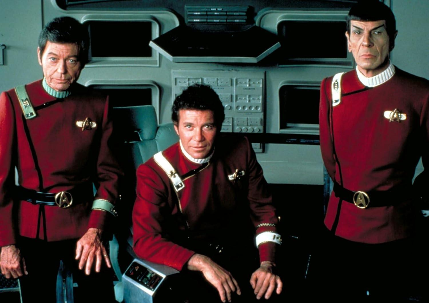 Drei Männer in roten Uniformen von Star Trek. Sie befinden sich in einem Kontrollraum des Schiffs. Der Mann in der Mitte sitzt.