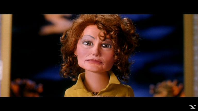 Eine Puppenfrau mit roten Locken und gelbem Hemd. Sie hat einen strengen Ausdruck und blickt in die Kamera. Dunkelblauer Hintergrund.