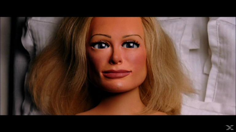 Nahaufnahme eines Puppengesichts mit blonden Haaren, blauen Augen und rosigen Wangen. Die Puppe liegt auf einer weißen Oberfläche.