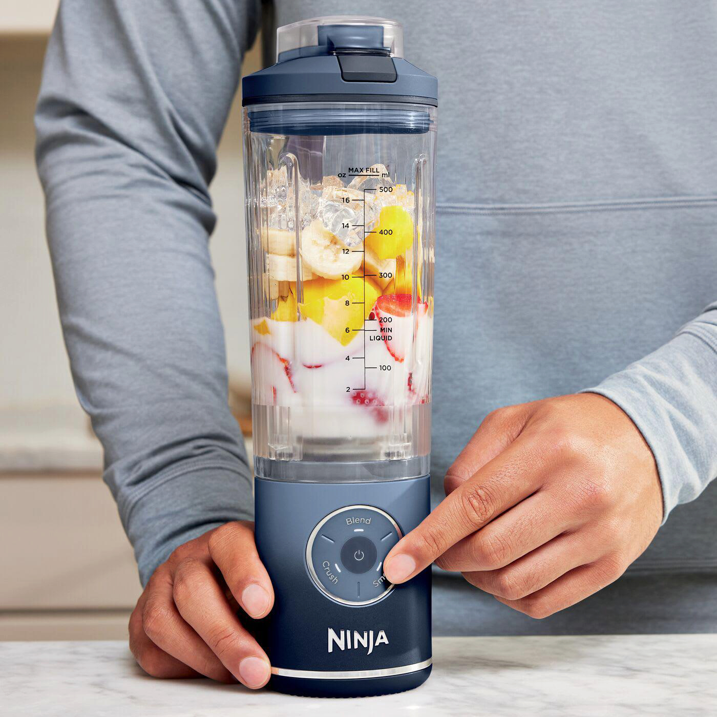 Eine Person benutzt einen Ninja-Mixer. Der Mixer ist mit Früchten und Eis gefüllt. Die Person drückt die Starttaste.
