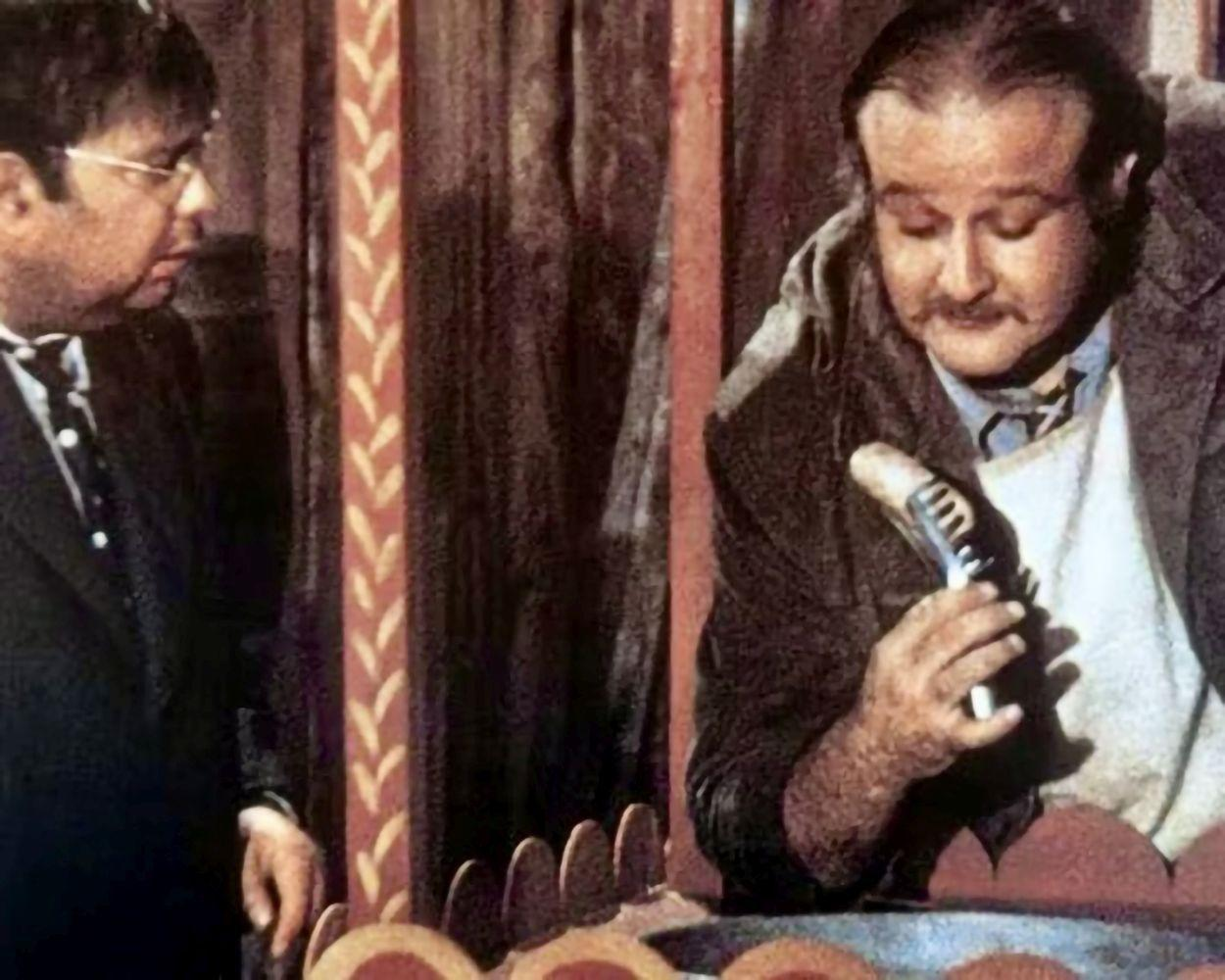 Zwei Männer in einer Szene. Einer hält einen Gegenstand, möglicherweise eine Wurst, der andere schaut zu. Verziertes Holzgerüst im Hintergrund.