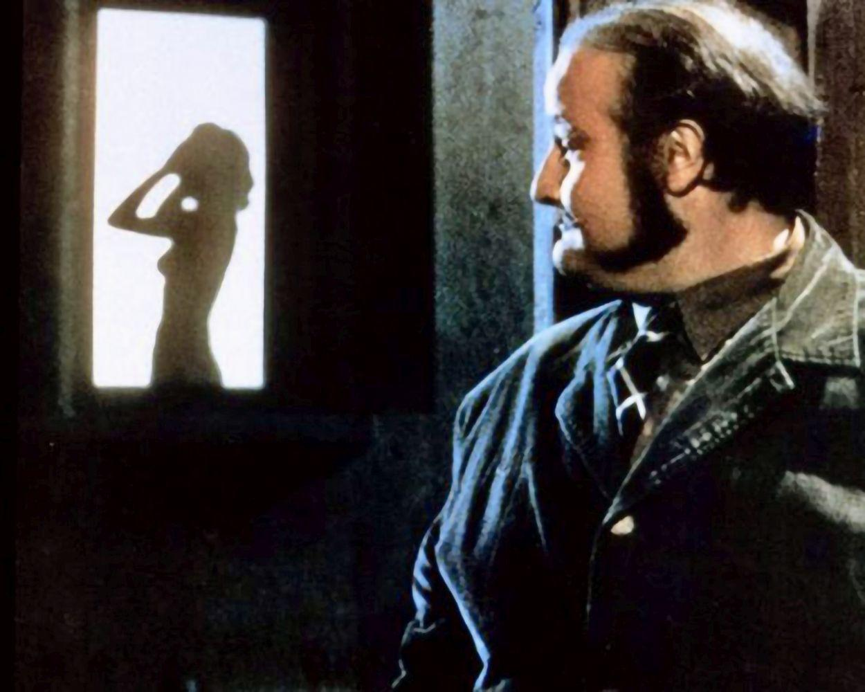 Ein Mann betrachtet die Silhouette einer Frau in einem hellen Fenster. Der Mann trägt eine Jeansjacke und hat einen Bart.