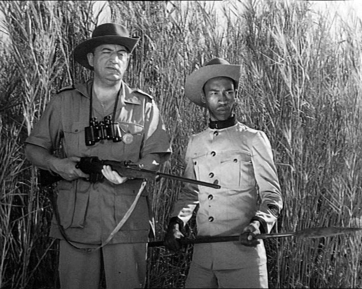Zwei Männer mit Hüten stehen in hohem Gras. Einer hält ein Gewehr, der andere einen Speer. Beide tragen Uniformen, was auf einen historischen Kontext hindeutet.