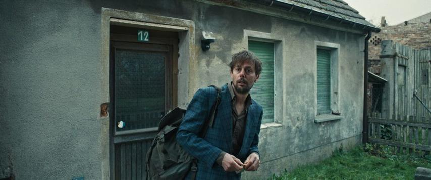 Ein Mann steht vor einem verwitterten Gebäude. Er trägt eine blaue Jacke und einen Rucksack. Die Nummer 12 steht auf der Tür.