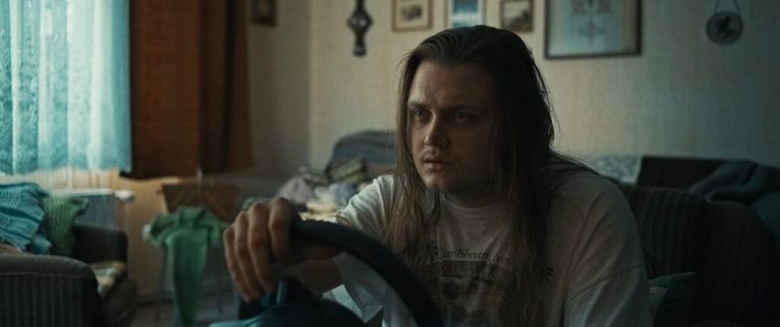 Ein Mann mit langen Haaren hält ein Lenkrad. Er ist in einem Raum mit einem Fenster und Bildern an der Wand und wirkt besorgt.