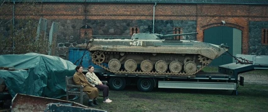 Zwei Frauen sitzen auf einer Bank neben einem Panzer auf einem Anhänger. Im Hintergrund ein Backsteingebäude.