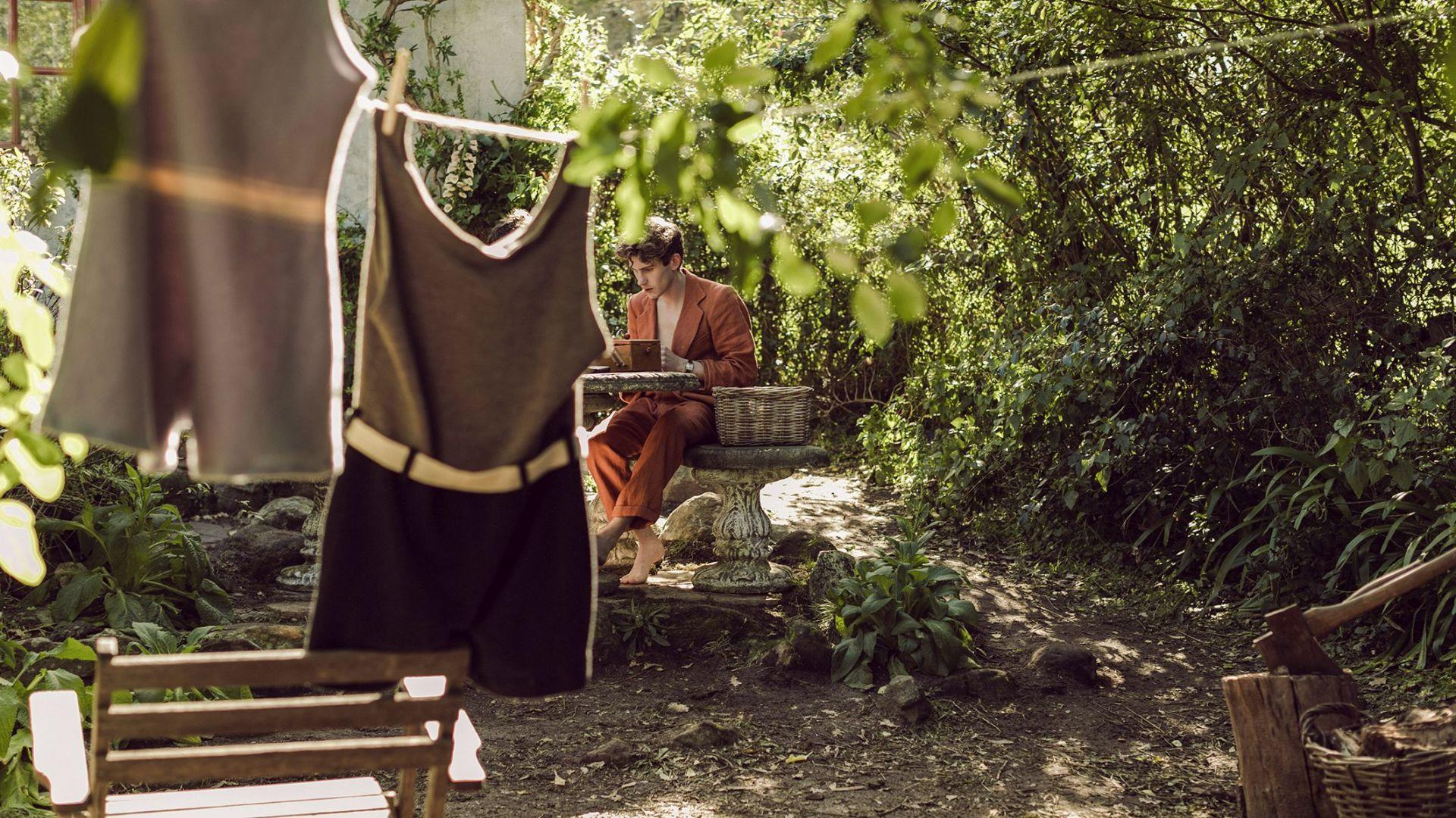Ein Mann in orangefarbenem Anzug sitzt in einem Garten an einem Tisch. Kleidung hängt im Vordergrund. Grünes Laub umgibt ihn.