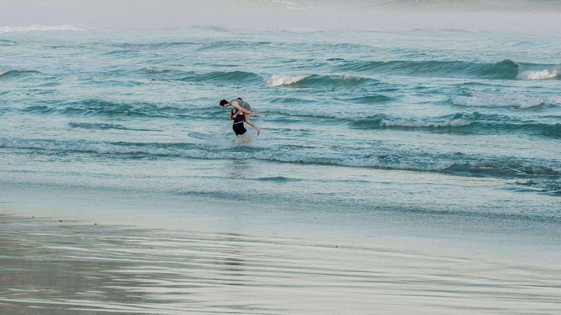 Zwei Personen im Meer. Einer trägt den anderen. Wellen im Hintergrund. Strand im Vordergrund. Bewölkter Himmel.