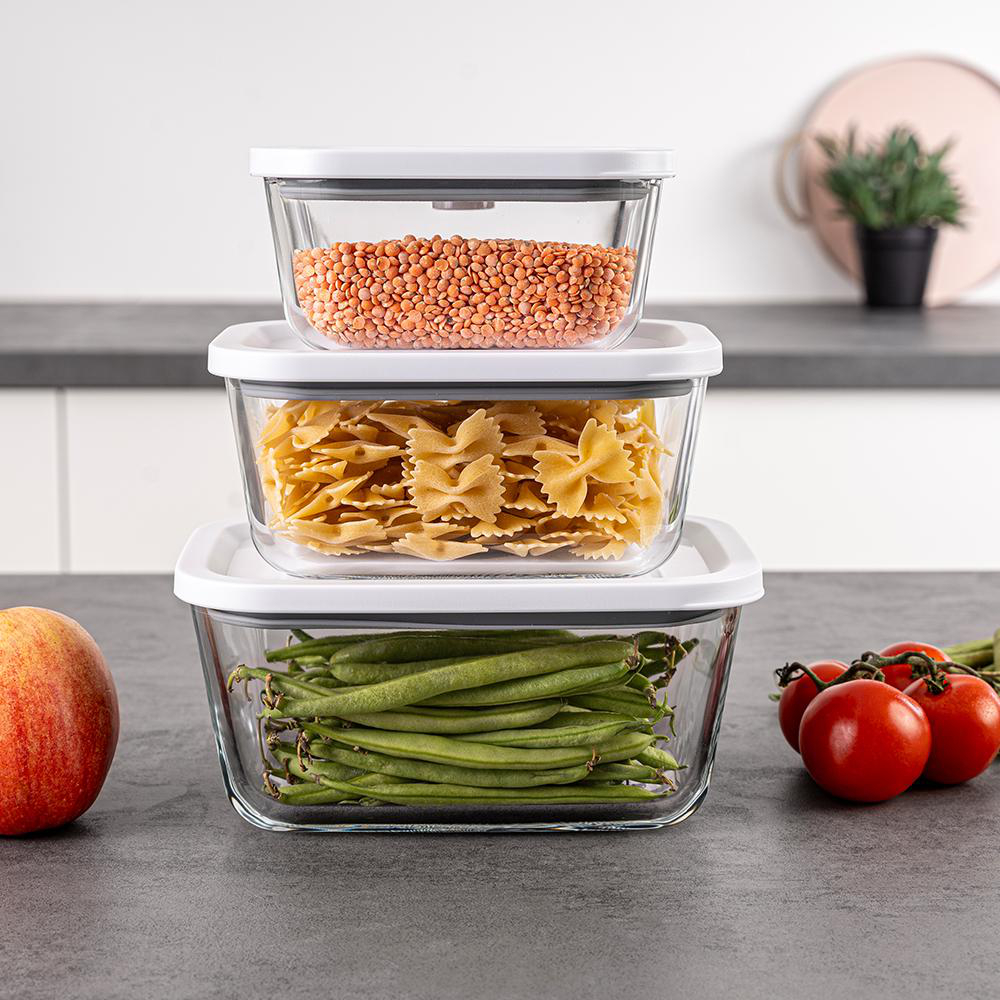 Drei gestapelte Glasbehälter, gefüllt mit Linsen, Pasta und grünen Bohnen, mit Tomaten und einem Apfel daneben.