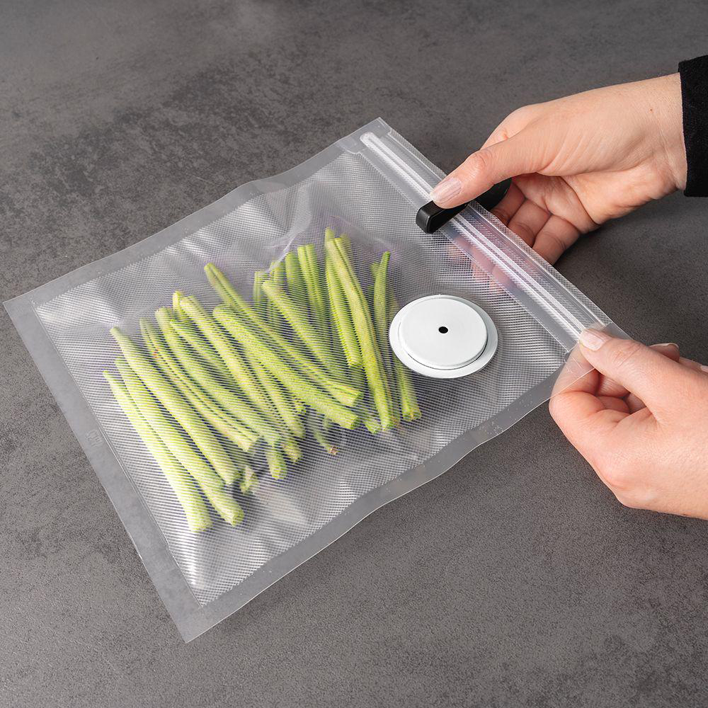 Hände verschließen einen Plastikbeutel mit grünen Bohnen. Eine weiße Scheibe mit schwarzem Punkt ist im Beutel.
