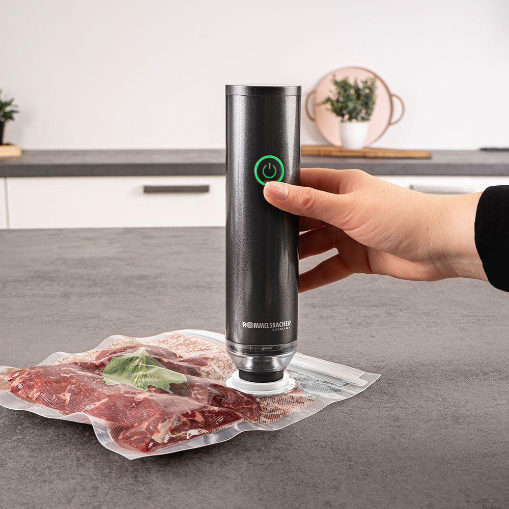 Eine Person benutzt einen Vakuumierer auf einem Beutel mit Fleisch. Das Gerät ist schwarz mit einem grünen Einschaltknopf. Im Hintergrund ist eine Küchenzeile.
