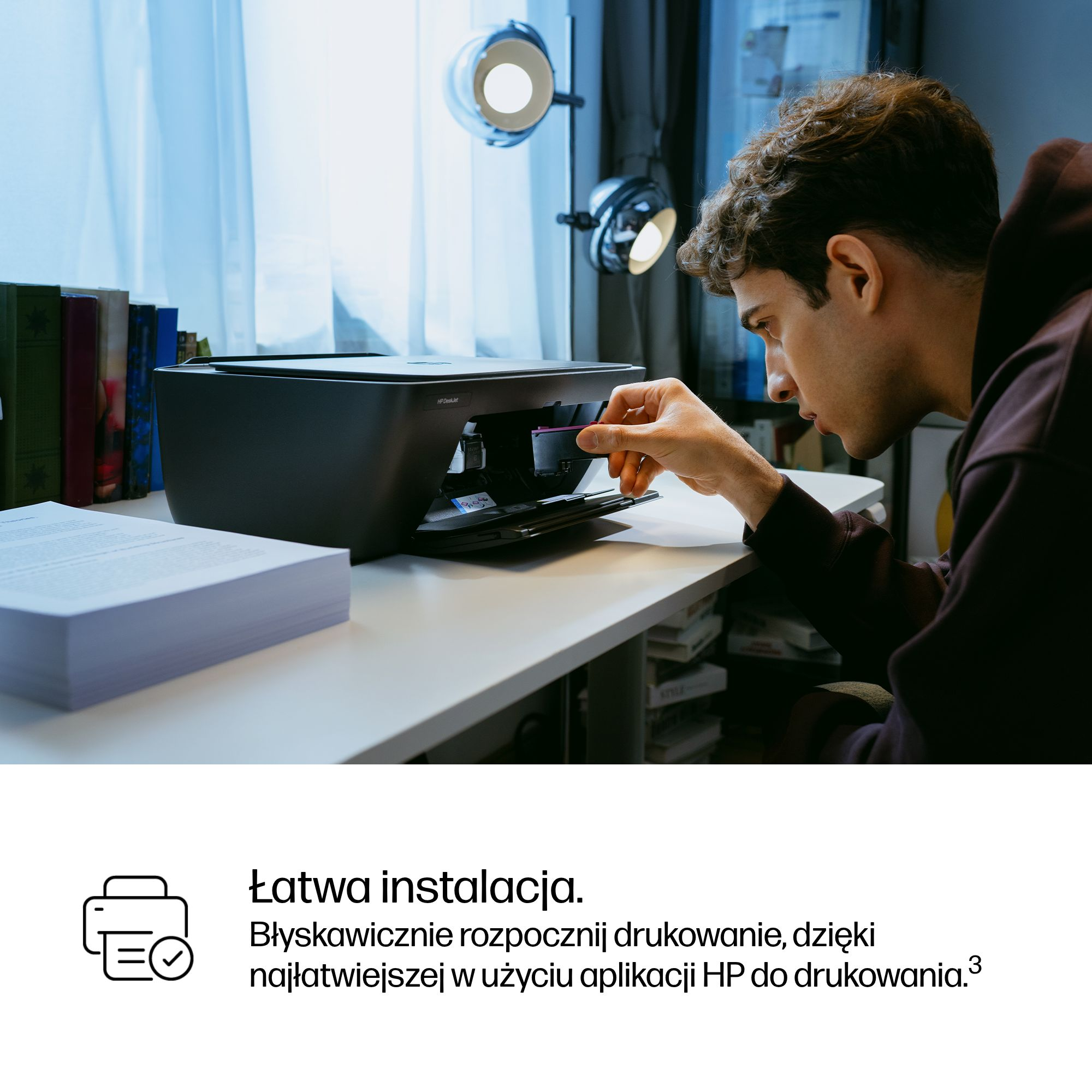 Mężczyzna instaluje wkład atramentowy w drukarce na biurku. Widoczne są książki i stos papieru.