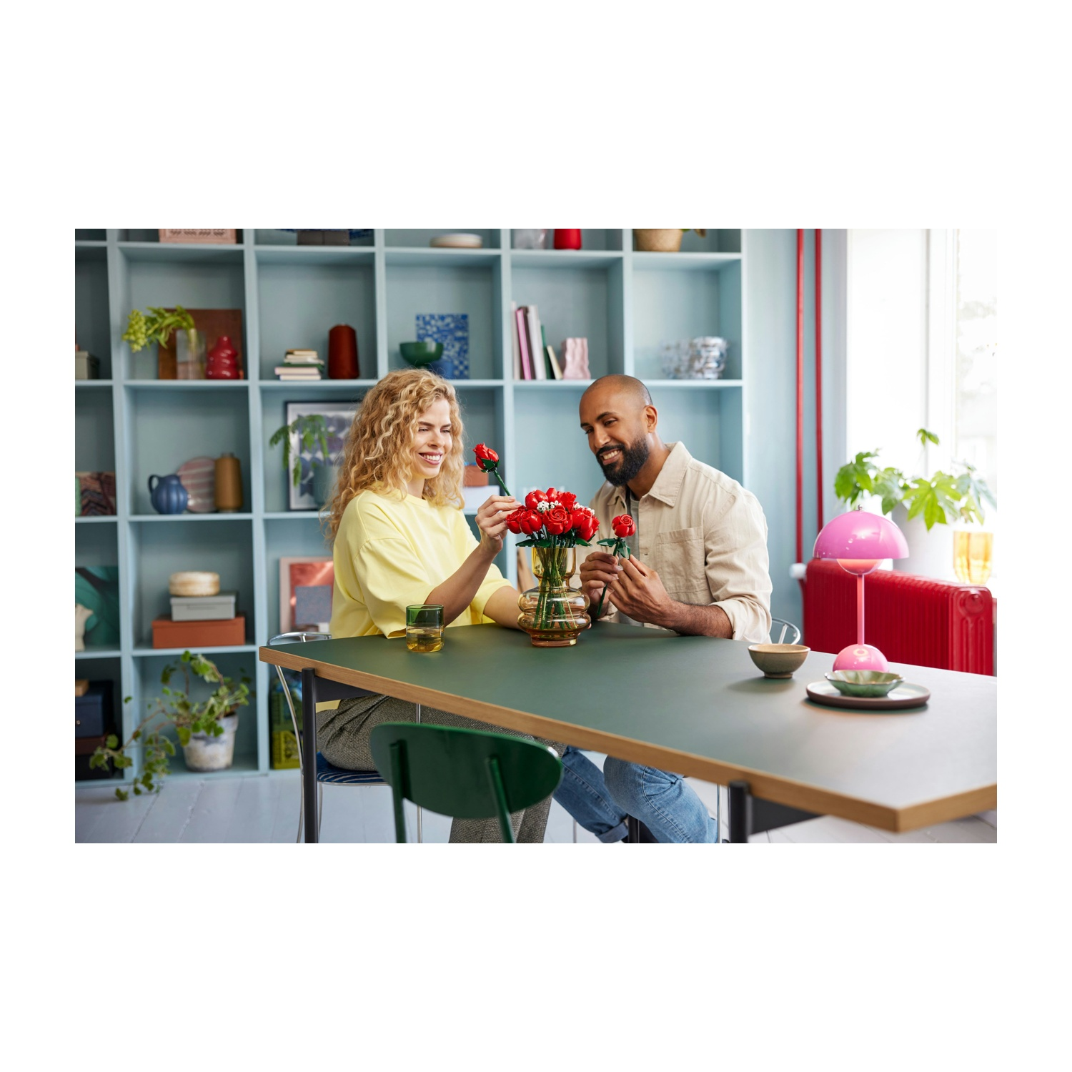 Una coppia sistema rose rosse in un vaso su un tavolo. Una lampada rosa e un radiatore rosso sullo sfondo.